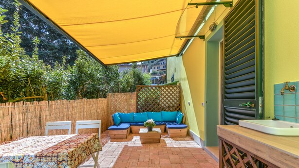 Eine Terrasse mit gelber Markise, einem blauen Sofa, einem Tisch mit gemustertem Tischläufer und einer Holzbank.