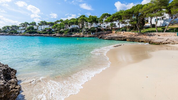 A beach with clear turquoise water, sandy shore, and white buildings with trees. - Apartment 'White Suites' with Shared Pool, Wi-Fi and Air Conditioning (Cala D'or)