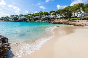 A beach with clear turquoise water, sandy shore, and white buildings with trees. - Apartment 'White Suites 3' with Shared Pool, Wi-Fi and Air Conditioning (Cala D'or)