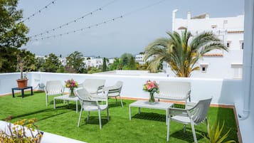Eine Dachterrasse mit weißer Gartenmöbel, einem kleinen Tisch mit Blumenvase und Lichterketten.