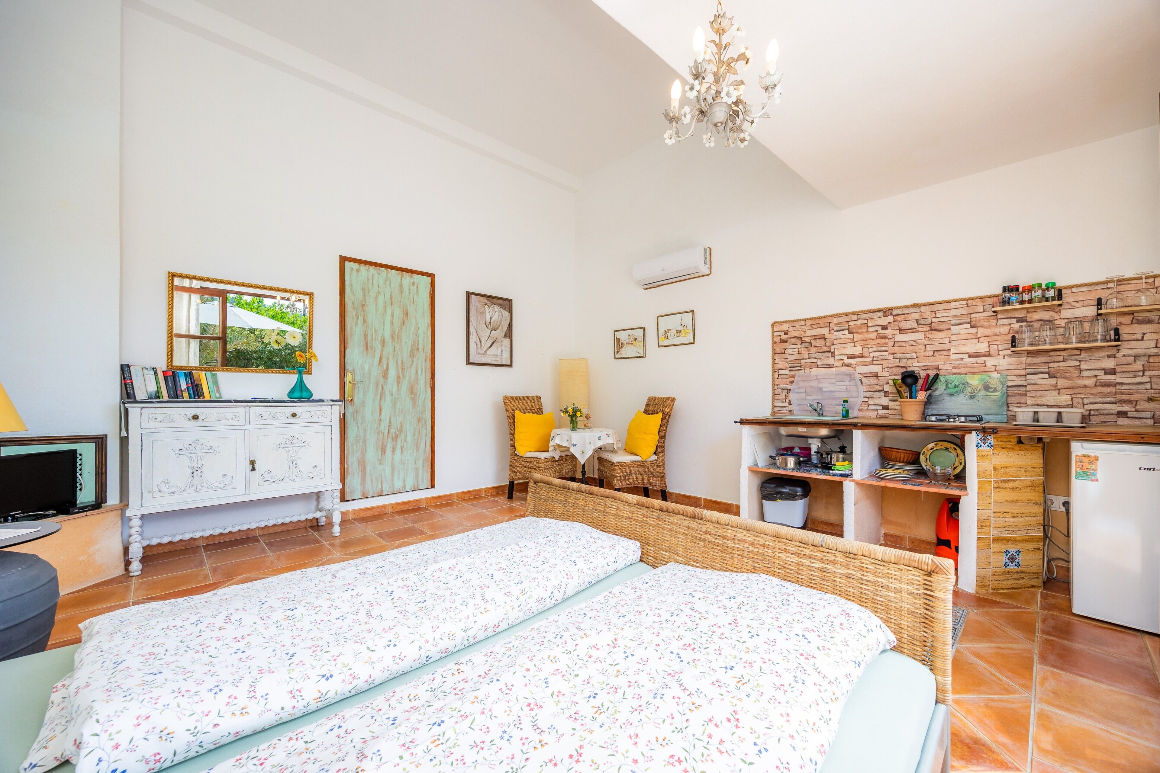 A bedroom with two beds, a dresser, a television, a dining area with a table and chairs, and a kitchen with a refrigerator and shelves.