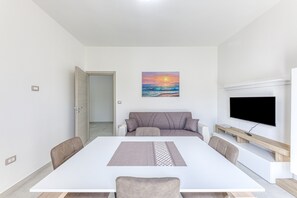 A modern living room with a white sofa, a flat-screen TV, a painting of a sunset, and a dining table with chairs.
