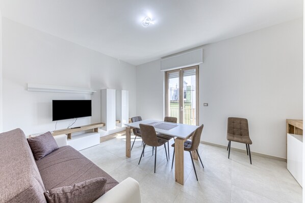 En moderne stue med et fladskærms-tv, et spisebord med stole, en sofa og et vindue med persienner.