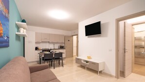 A modern living room with a sofa, a dining area with chairs, a kitchen with cabinets, a TV mounted on the wall, and a door leading to another room. - Holiday Apartment 'Appartamento A Pigna' with Balcony, Wi-Fi and Air Conditioning (Balestrate)