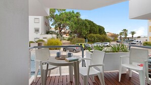 A balcony with white wicker furniture, a table with a book and two cups, and a view of a street with cars and trees. - Apartment 'Golden Ocean' with Shared Pool, Wi-Fi and Air Conditioning (Cabanas de Tavira)