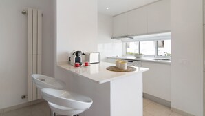 A modern kitchen with white cabinetry, a central island with bar stools, a toaster, and a coffee maker. - Apartment 'Golden Ocean' with Shared Pool, Wi-Fi and Air Conditioning (Cabanas de Tavira)