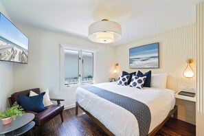 A hotel room with a large bed, a desk with a chair, a small table with a plant, a window with a view of the beach, and a painting on the wall. - Newly Renovated Beach Cottage in Seaside | King Bed | 194 Ft to the Beach (Santa Rosa Beach)