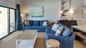 Ein modernes Wohnzimmer mit einer blauen Sofagarnitur, einem Holztisch und einem Esstisch. An der Wand hängt ein Gemälde mit Segelbooten, eine Deckenflatter sorgt für Luftzirkulation und ein großes Fenster ist mit Vorhängen versehen.