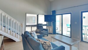 Ein modernes Wohnzimmer mit einem blauen Sofa, einem Couchtisch und einem Fernseher. Eine Treppe führt zu einem Balkon mit Gartenmöbeln.