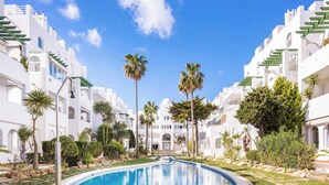 Een zwembad omringd door witte gebouwen met balkons, palmbomen en een helderblauwe lucht.