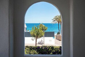 A view through an arched window of a coastal area with a clear blue sea, palm trees, and a building in the background. - Cairo Apartment (Jávea)