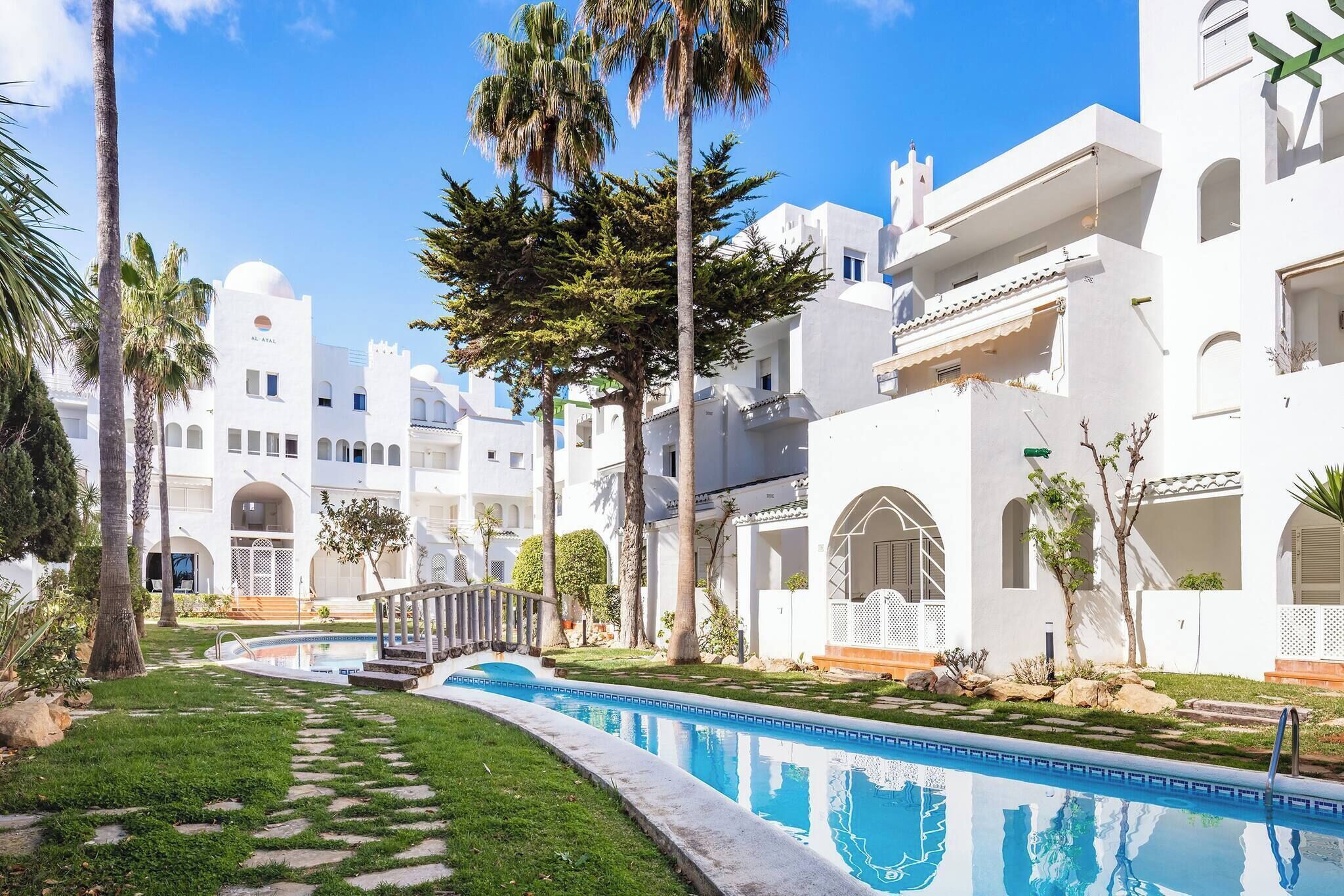 A pool area with a clear blue pool, surrounded by white buildings with arched windows and balconies, palm trees, and a well-maintained lawn.