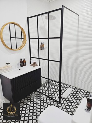 Une salle de bain avec un sol carrelé en noir et blanc, un lavabo blanc avec un meuble noir, un miroir encadré en bois et une cabine de douche noire.