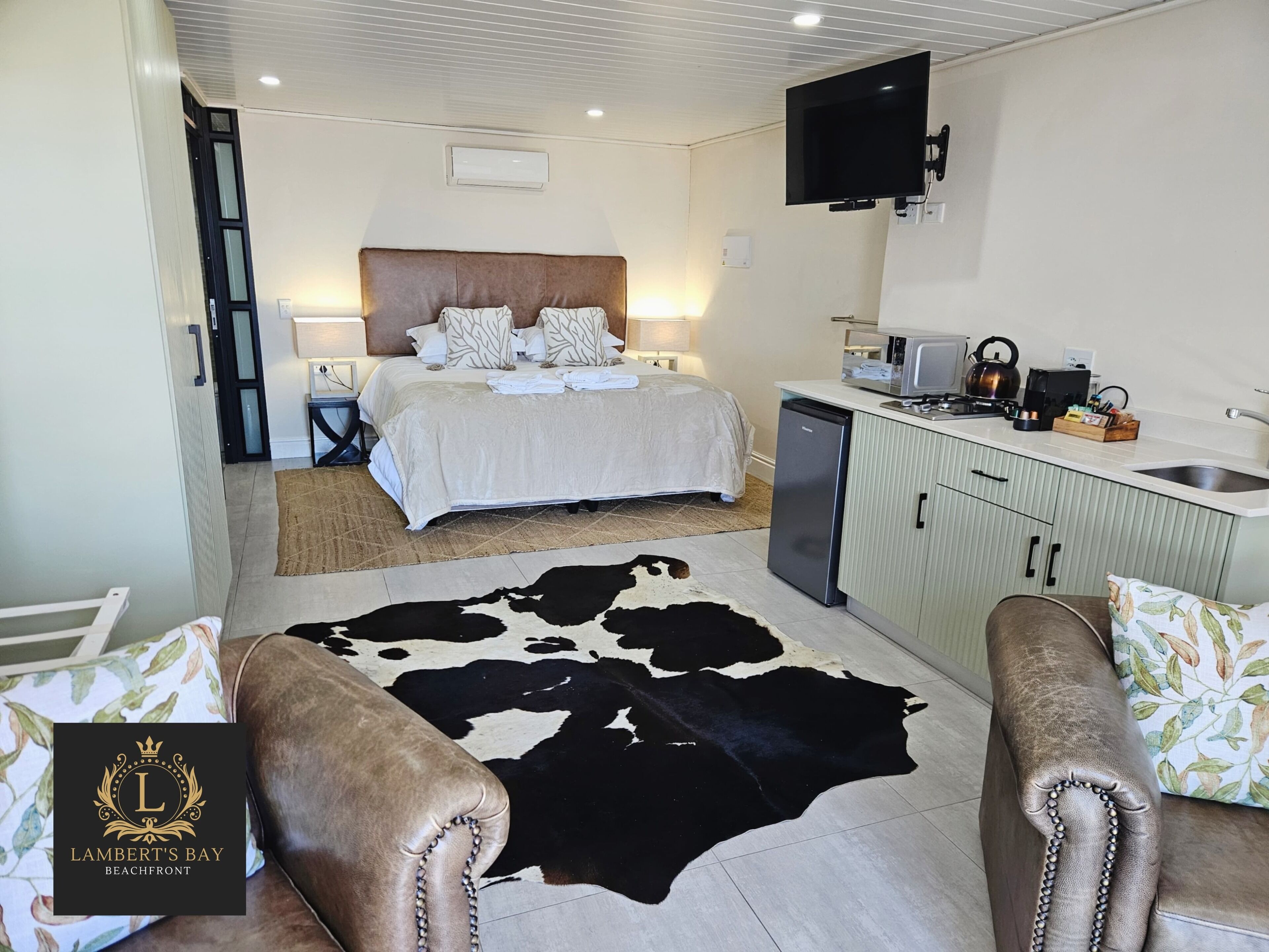 A hotel room with a bed, a kitchen area, a flat-screen TV, and a cowhide rug.