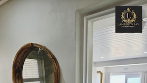 A bathroom with a round mirror, sink, and toiletries. A bedroom with a bed, towels, and a view of the beach is visible through an open door. - Lambert's Bay Beachfront (Lambert's Bay)