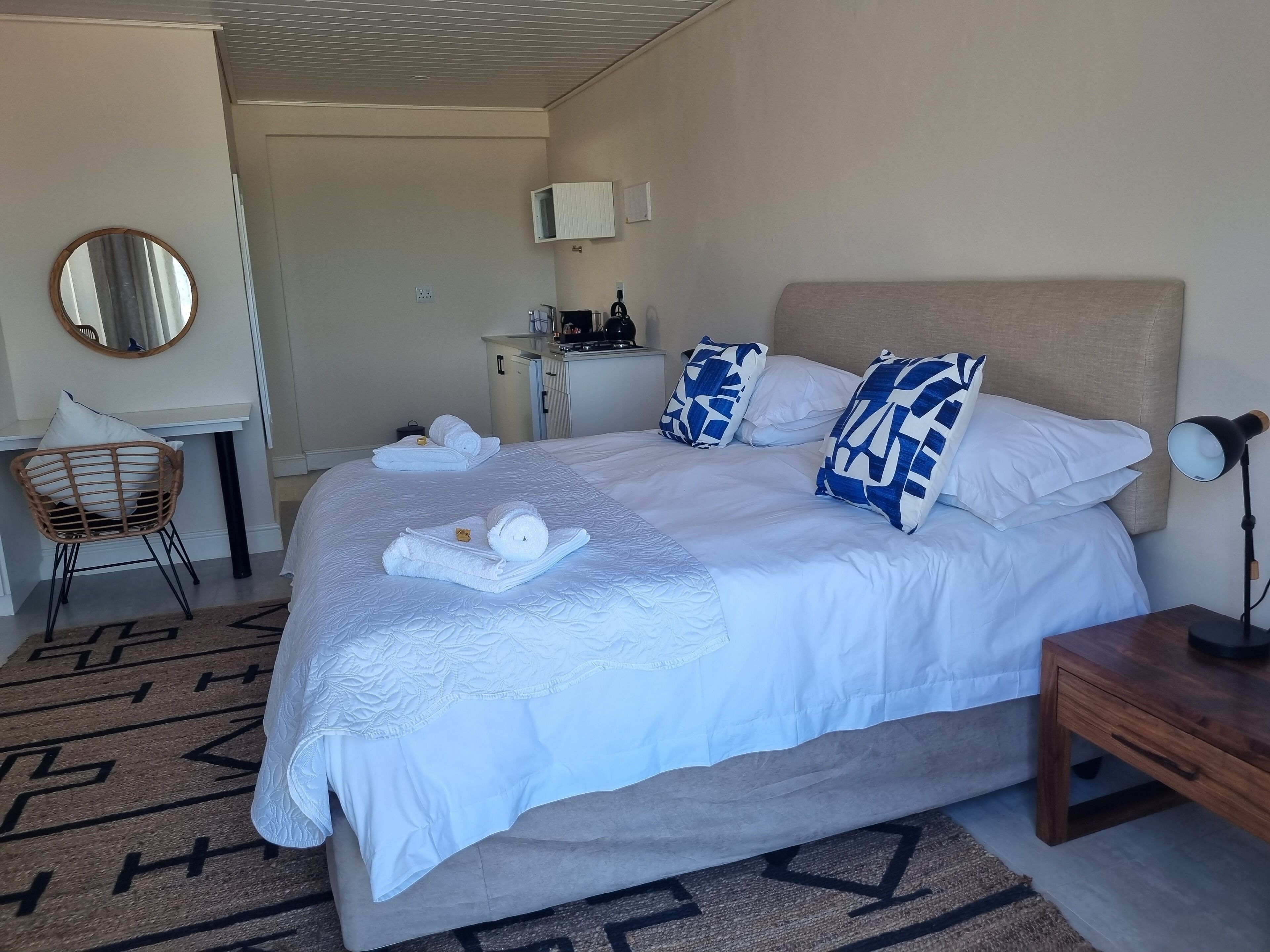 A neatly made bed with white linens and blue patterned pillows, a bedside table with a lamp, and a chair with a woven backrest.