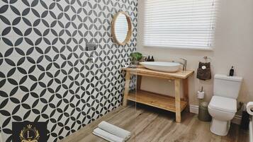 A bathroom with a geometric tile pattern, a wooden vanity, a sink, a toilet, and a window with blinds.