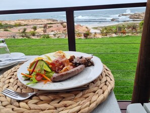 Un repas servi sur un tapis tressé, avec vue sur une plage rocheuse et l’océan.
