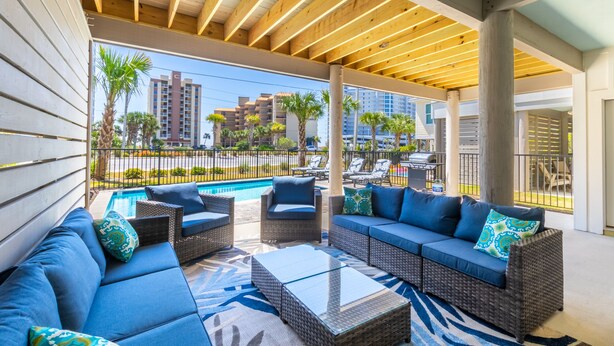 A covered outdoor seating area with wicker furniture and a pool in the background. - Luxury 6-Bedroom Retreat | Pool & Beach Access (Gulf Shores)