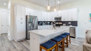 A modern kitchen with white cabinets, stainless steel appliances, a central island with bar stools, and pendant lighting. - Luxury 6-Bedroom Retreat | Pool & Beach Access (Gulf Shores)