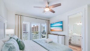 A bedroom with a bed, a ceiling fan, a TV, and a view of the beach through the window. - Luxury 6-Bedroom Retreat | Pool & Beach Access (Gulf Shores)
