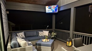 A covered outdoor seating area with a sofa, ottoman, and wicker chairs. A TV is mounted on the ceiling, and a lantern is on the floor. - Spacious mountain getaway on golf course, near trails, waterfalls & breweries! (Laurel Park)