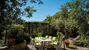 A patio area with a round table and green cushioned chairs, surrounded by lush greenery and potted plants.