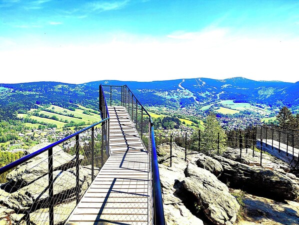 A wooden suspension bridge with metal railings, leading over a rocky terrain with a view of a valley and distant mountains.