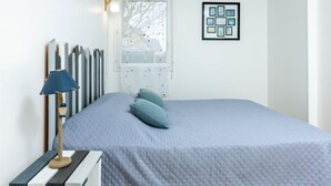 A bedroom with a bed, a nightstand, a lamp, a framed picture, and a window with blinds. - Les Caps Horniers by Interhome (Pleurtuit)