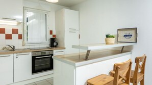 A modern kitchen with white cabinets, a built-in oven, a coffee maker, and a small wooden table with chairs. - Les Caps Horniers by Interhome (Pleurtuit)
