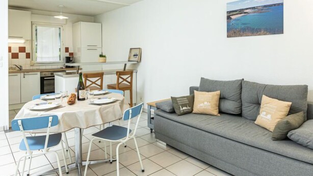 A compact living space with a dining area, a grey sofa with decorative pillows, and a coastal view painting. - Les Caps Horniers by Interhome (Pleurtuit)