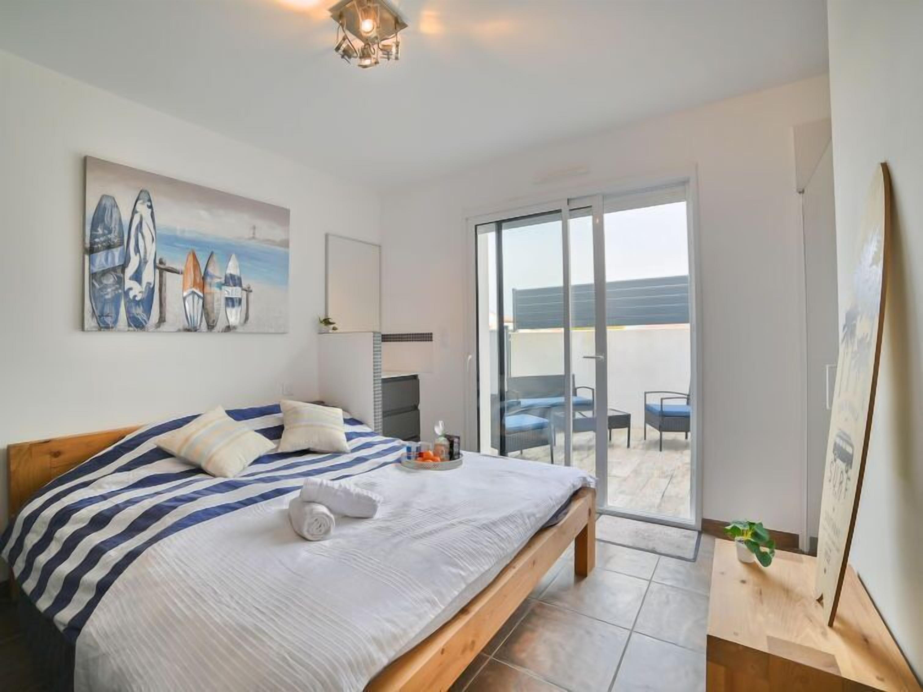 A bedroom with a bed, a painting of surfboards, a sliding glass door to a balcony, and a small table with a plant.