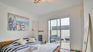 Una camera da letto con un letto, un quadro raffigurante tavole da surf, una porta scorrevole in vetro che dà su un balcone e un piccolo tavolo con una pianta.