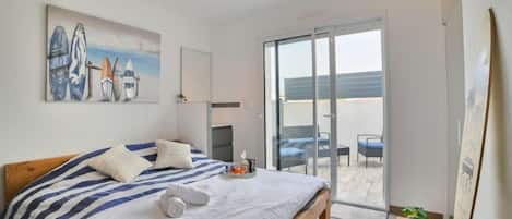 A bedroom with a bed, a painting of surfboards, a sliding glass door to a balcony, and a small table with a plant.