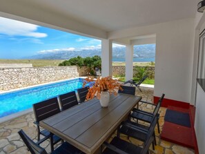 Ein Essbereich am Pool mit Tisch und Stühlen, eine Vase mit orangefarbenen Blumen und Blick auf Berge und Meer.