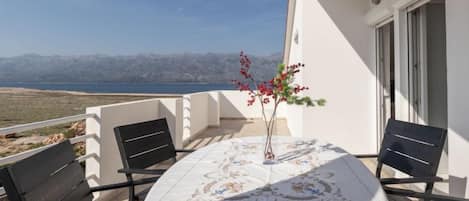 A balcony with a table set for two, a vase with flowers, and a view of the sea and mountains.