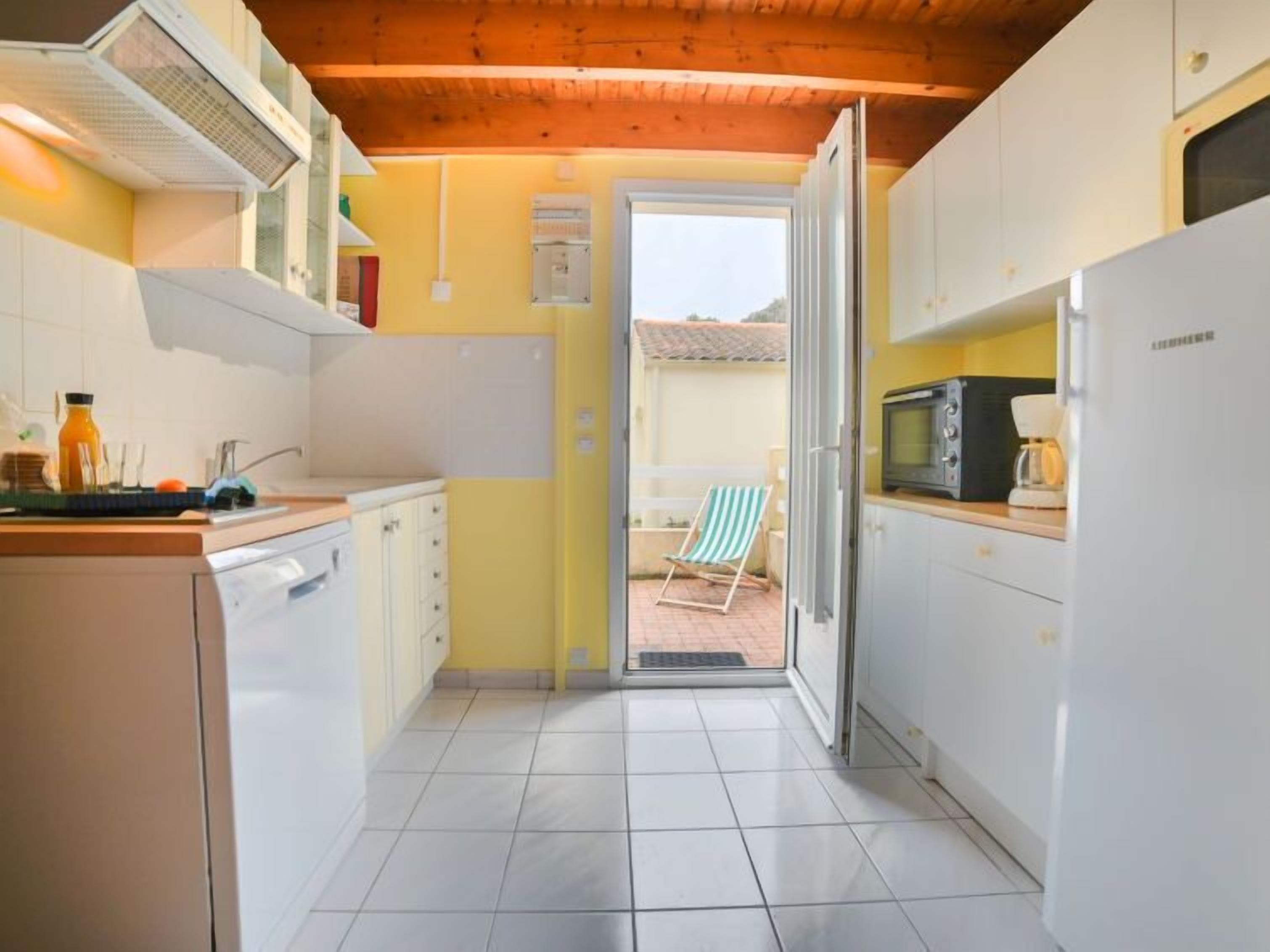 A compact kitchen with white cabinets, a tiled floor, and a wooden ceiling. A door leads to a balcony with a chair.