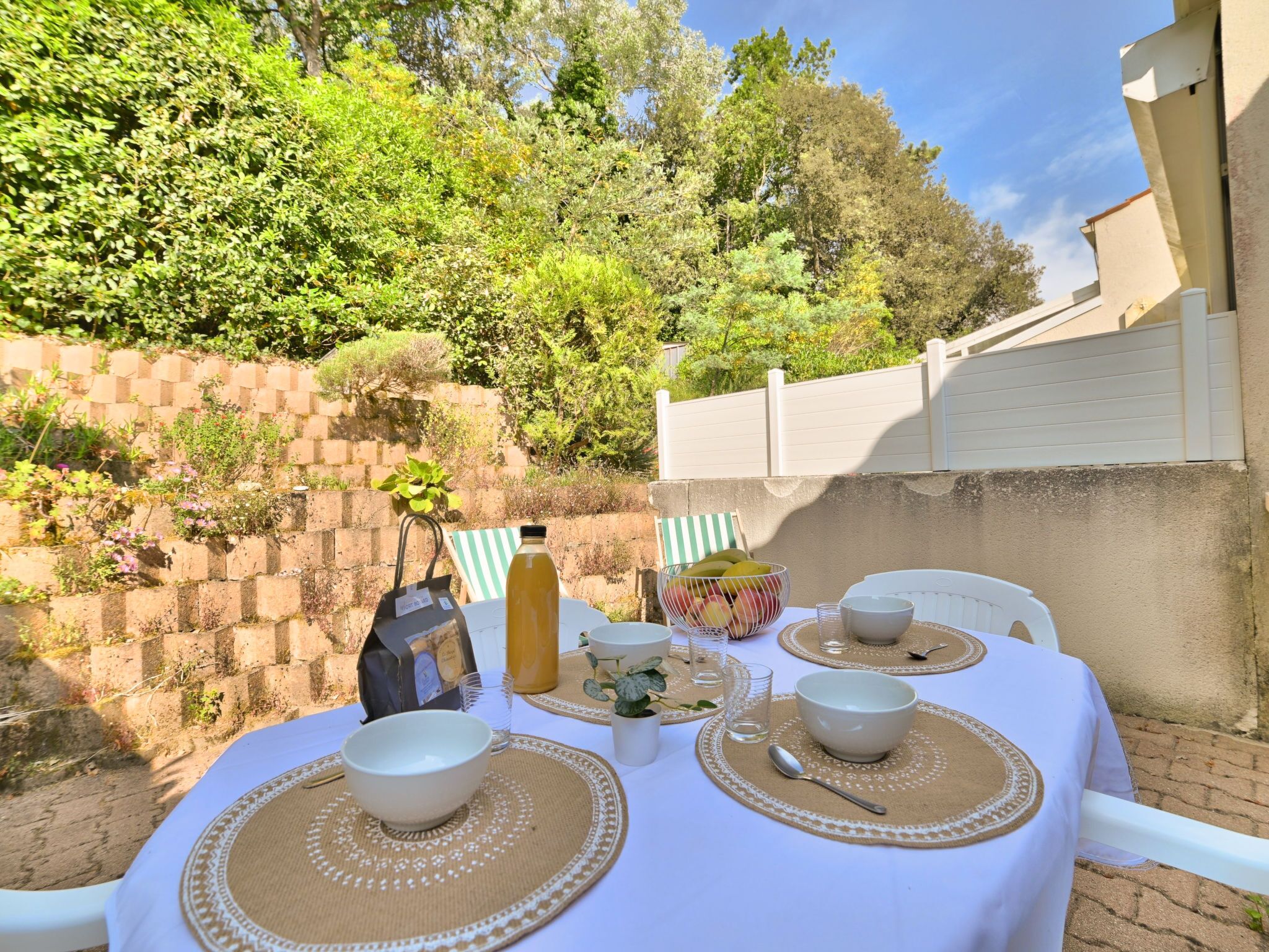 Un área de comedor al aire libre con una mesa preparada para dos personas, rodeada de vegetación y un muro de ladrillo.