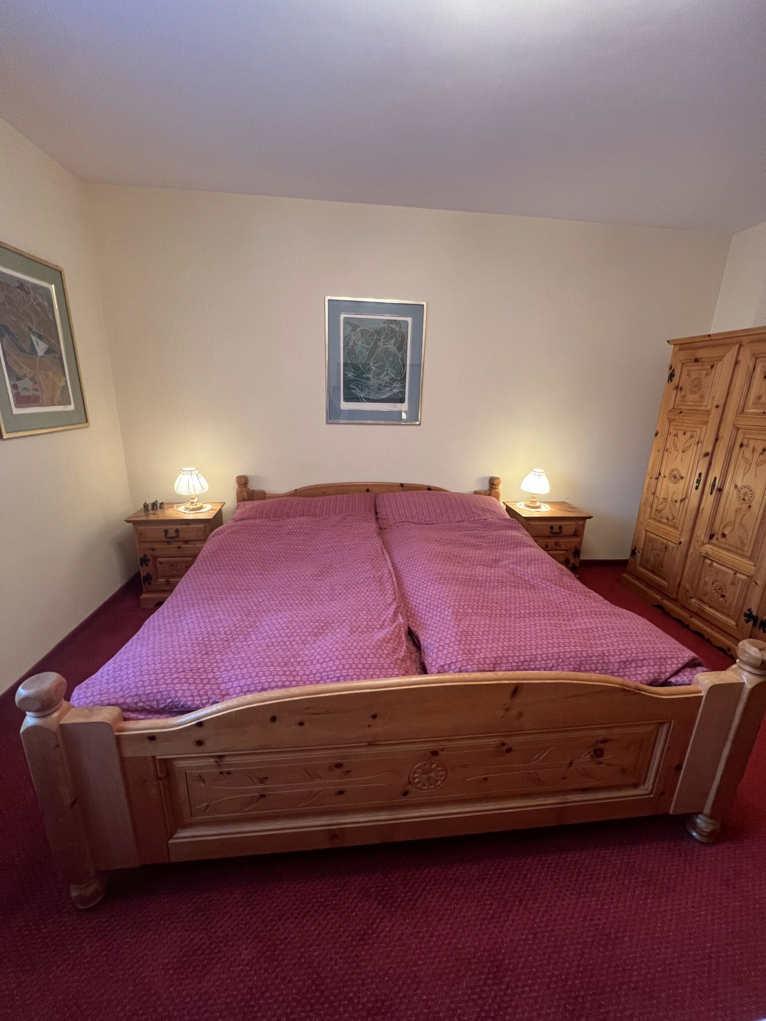 Un lit en bois surmonté d’une couvre-lit rose, dans une pièce dotée d’un tapis rouge, d’une armoire en bois et de deux tables de chevet avec des lampes.