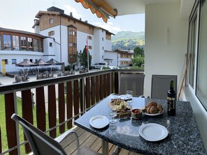 A balcony with a table set for dining, overlooking a mountainous landscape and a building with outdoor seating. - Haus Colani Ingold by Interhome (Valbella)