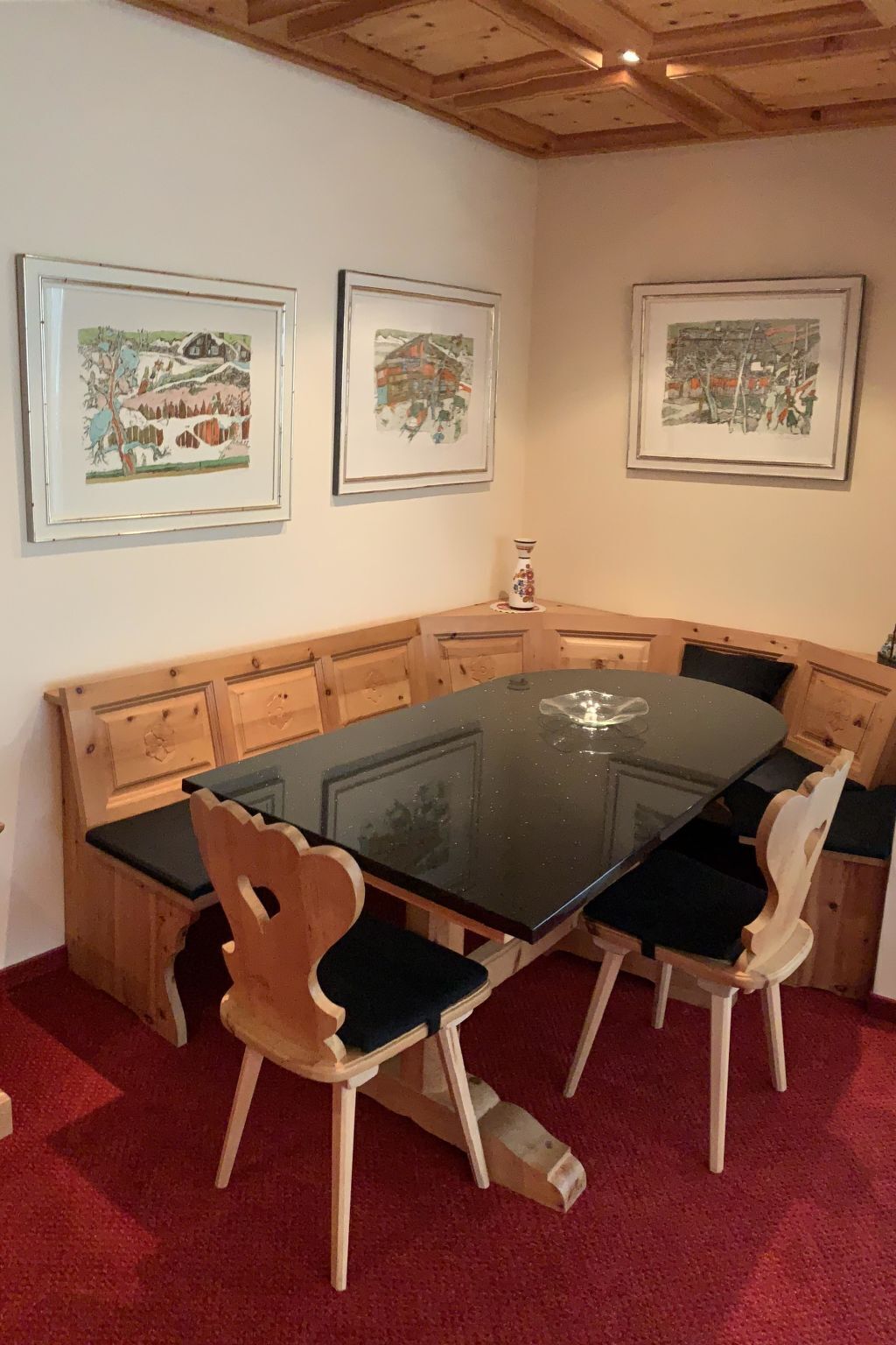 Ein Essbereich mit einem Tisch mit Glasplatte, umgeben von Holzstühlen mit gepolsterten Sitzen. An der Wand über dem Tisch hängen drei gerahmte Kunstwerke.