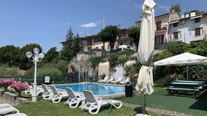 Una piscina con sillas de descanso y sombrillas, rodeada de vegetación y edificios.