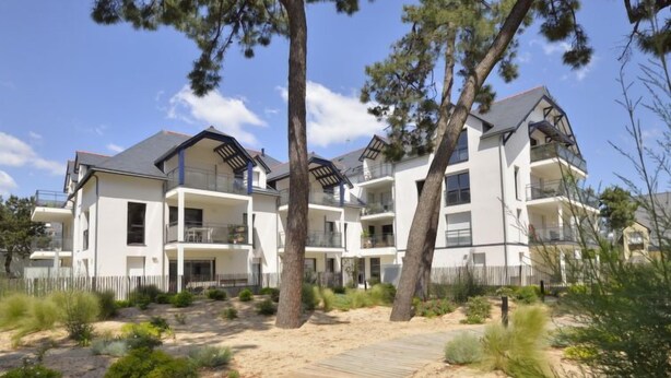 Un immeuble résidentiel moderne à plusieurs étages, doté de balcons, d’une terrasse en bois et entouré d’un paysage sablonneux.