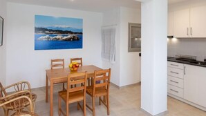 Un comedor con mesa y sillas, un cuadro de un paisaje costero, y una cocina con armarios blancos.