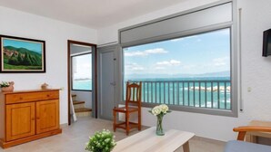 Una habitación con un ventanal grande que da a la costa, un armario de madera, una silla, una mesita con un florero y un cuadro en la pared.