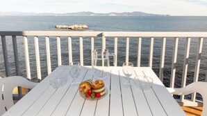 Una mesa blanca con un vaso de agua, dos vasos y un plato con comida y un bol en un balcón con vistas al mar.
