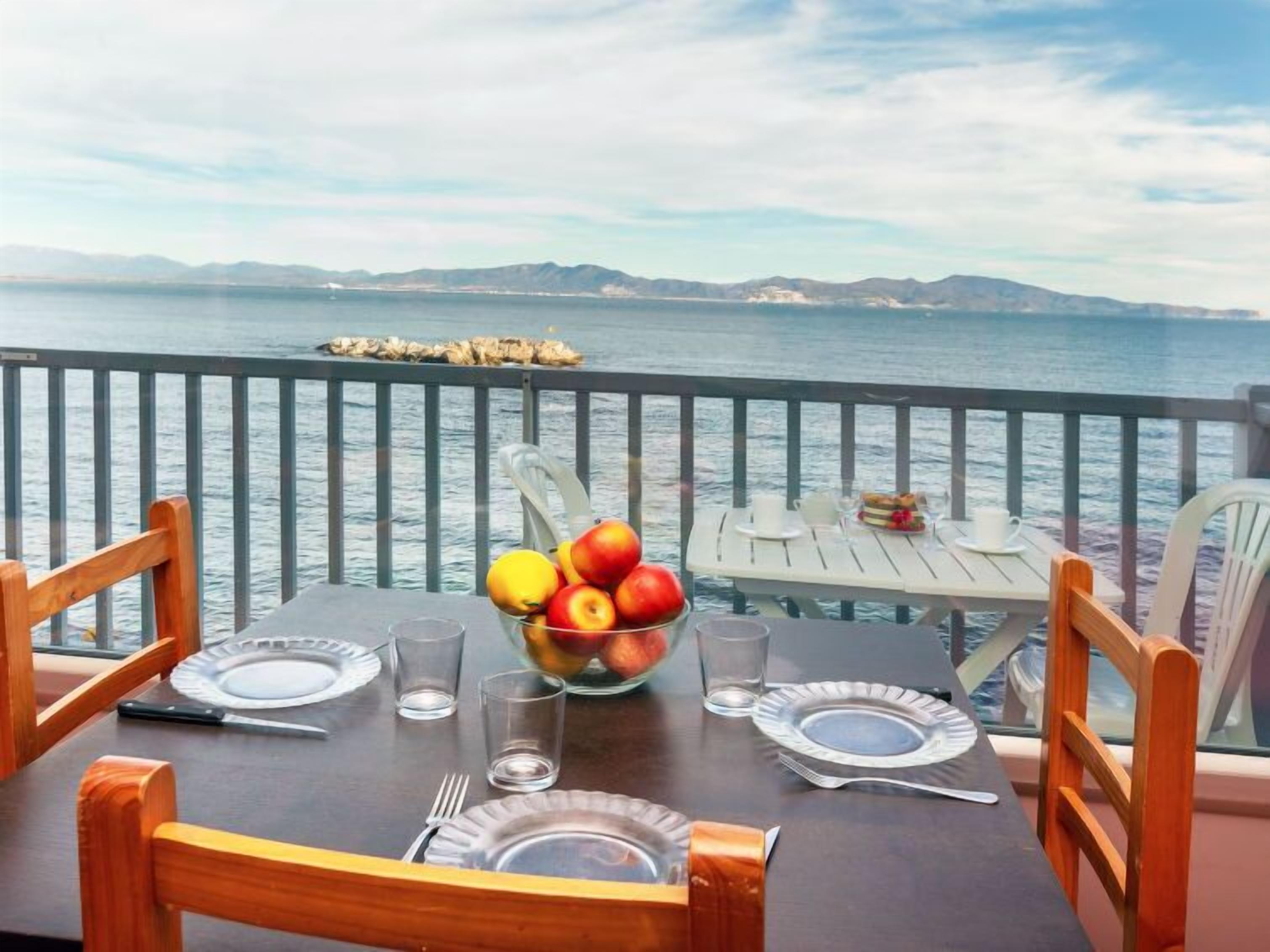 A table set for two with a fruit bowl, plates, and cutlery, overlooking a body of water with a distant shoreline.