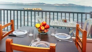 Une table dressée pour deux personnes, avec un panier de fruits, des assiettes et des couverts, donnant sur une étendue d’eau dont on aperçoit la côte au loin.