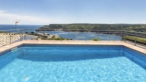 Ein klarer, blauer Pool mit dekorativem Rand, umgeben von einem Metallgeländer, mit Blick auf eine malerische Aussicht auf das Meer und die Hügel.