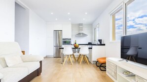 Una sala de estar moderna con sofá, un comedor con mesa y sillas, una cocina con refrigerador y estufa, y un televisor sobre un mueble.